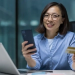 Smiling woman using smartphone and holding bank card at desk, representing easy online banking for expats with the top 10 banks in Germany