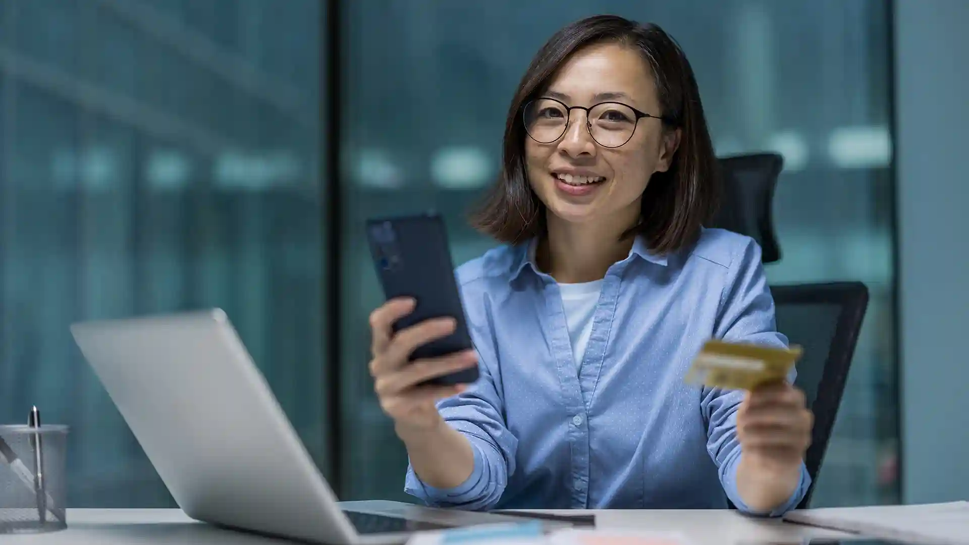 Smiling woman using smartphone and holding bank card at desk, representing easy online banking for expats with the top 10 banks in Germany